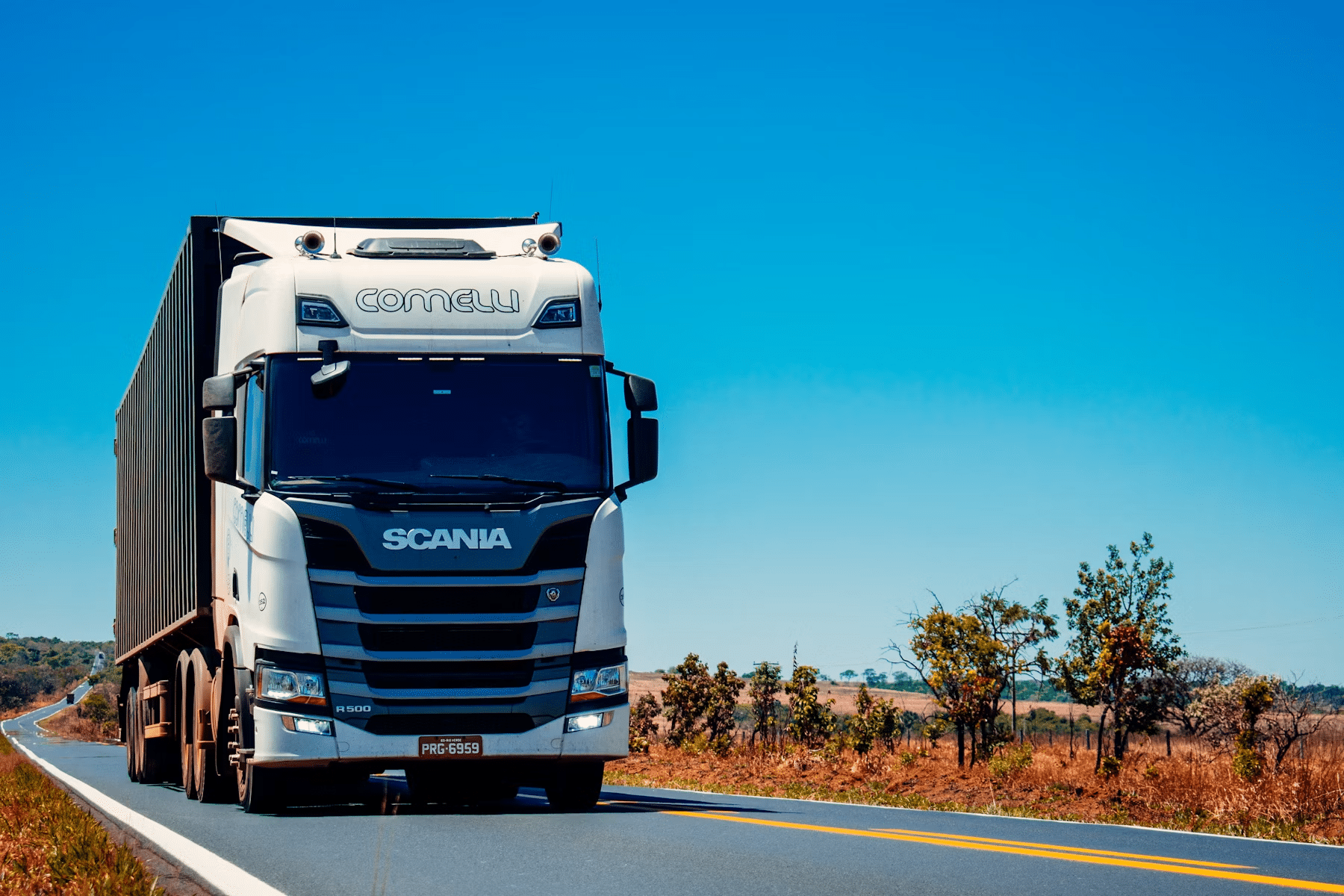 White Scania truck driving on a rural highway with clear blue skies in the background. White Scania truck driving on a rural highway with clear blue skies in the background.