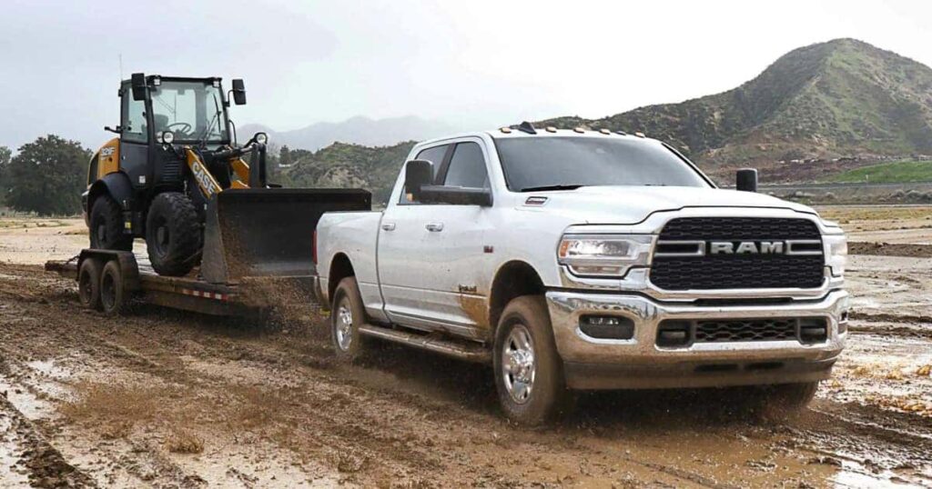 A white RAM truck tows a CASE construction loader on a muddy dirt road with mountains in the background.