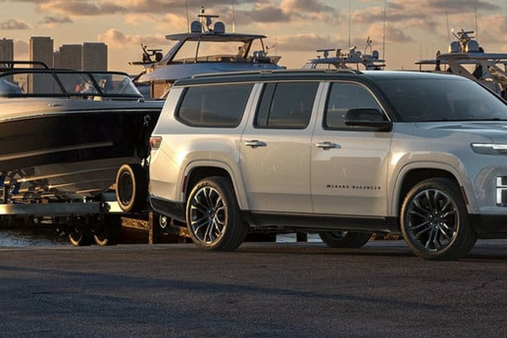 A white SUV towing a boat by a marina at sunset, with city skyline in the background.
